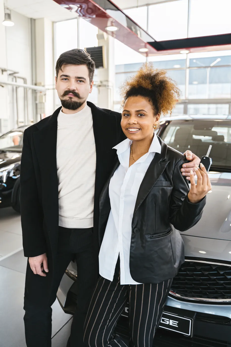 Smiling couple holding their new car keys beside the vehicle in a dealership showroom